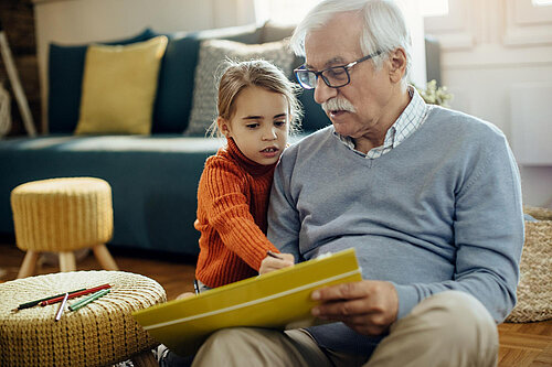 A young child is sat next to an older man, the child is pointing at a book held by the man